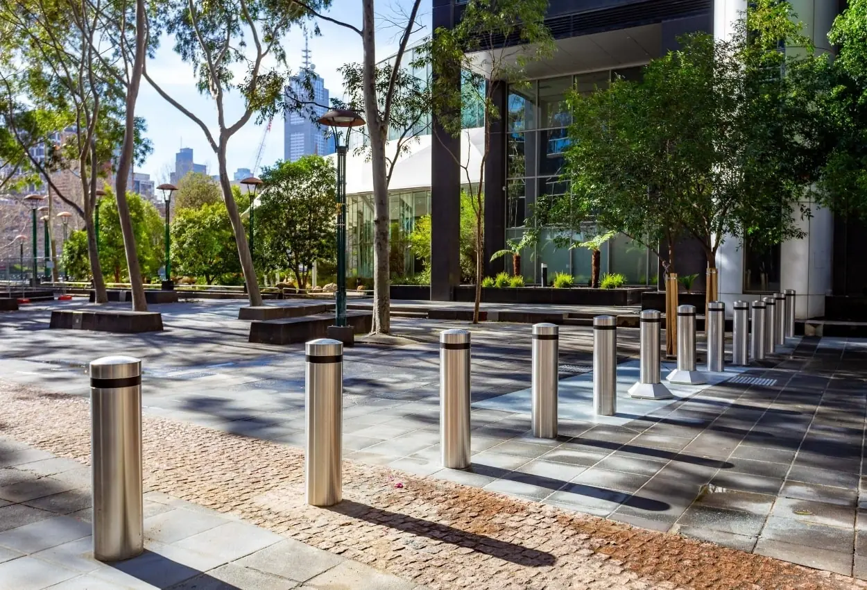 A row of stainless steel bollards lines a modern urban plaza in Australia with trees and buildings.