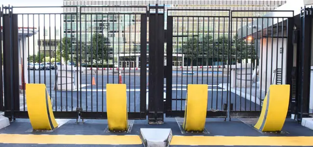 Four yellow Raptor bollards in front of a tall black security fence with a building complex in the background.