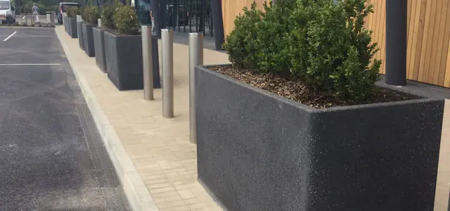 Concrete planters with greenery and metal bollards create a barrier along a pedestrian path.