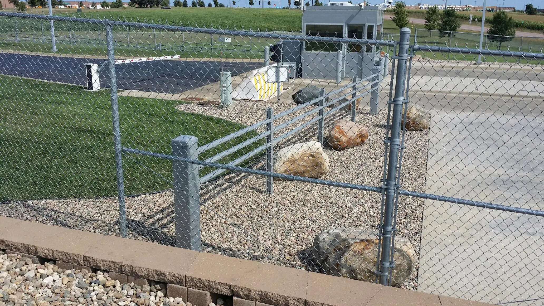 Chain-link fence with steel beams, rocks, and a guard booth in a secured area.